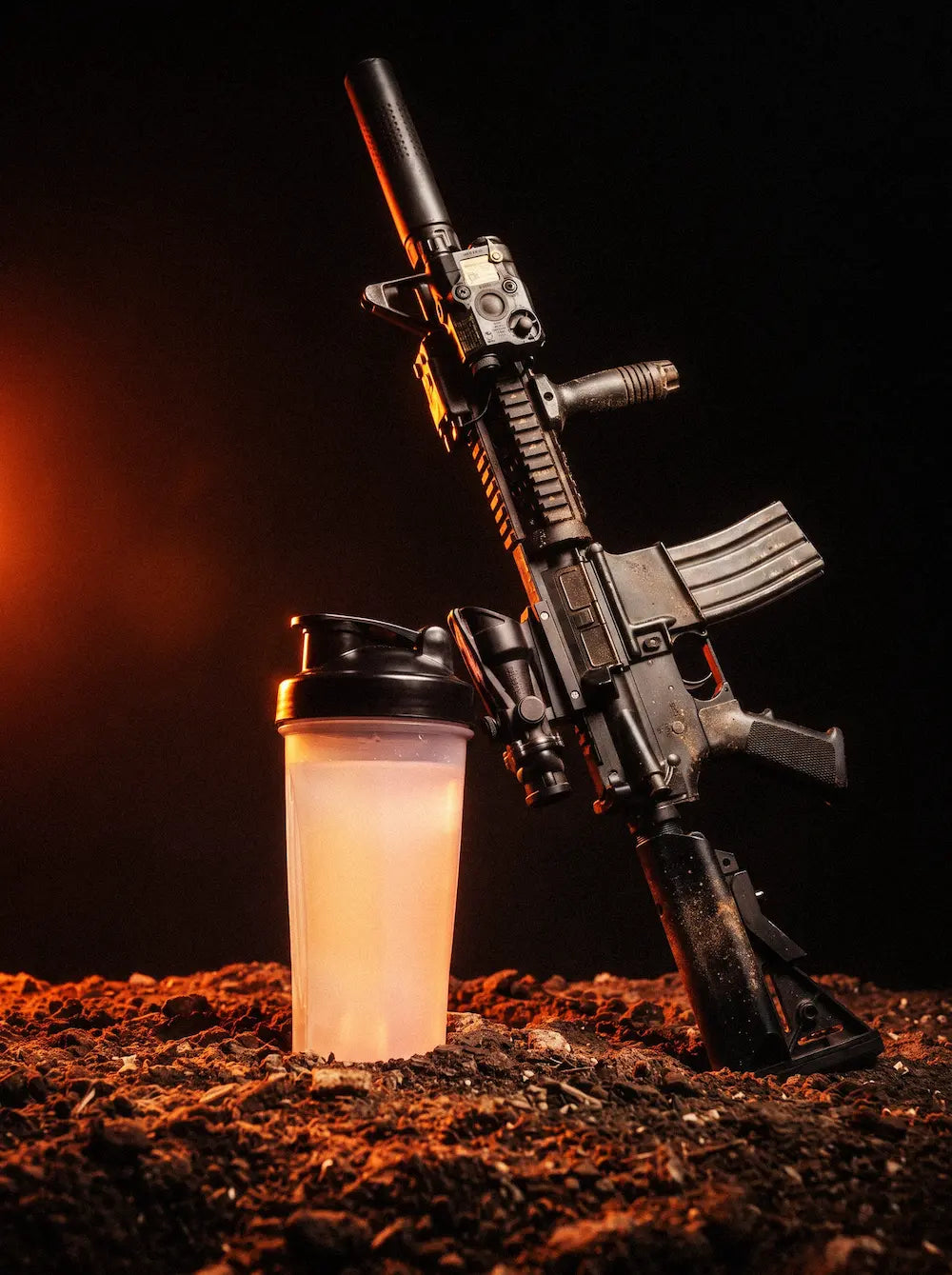Clear shaker with an orange drink beside a rifle on rocky ground.